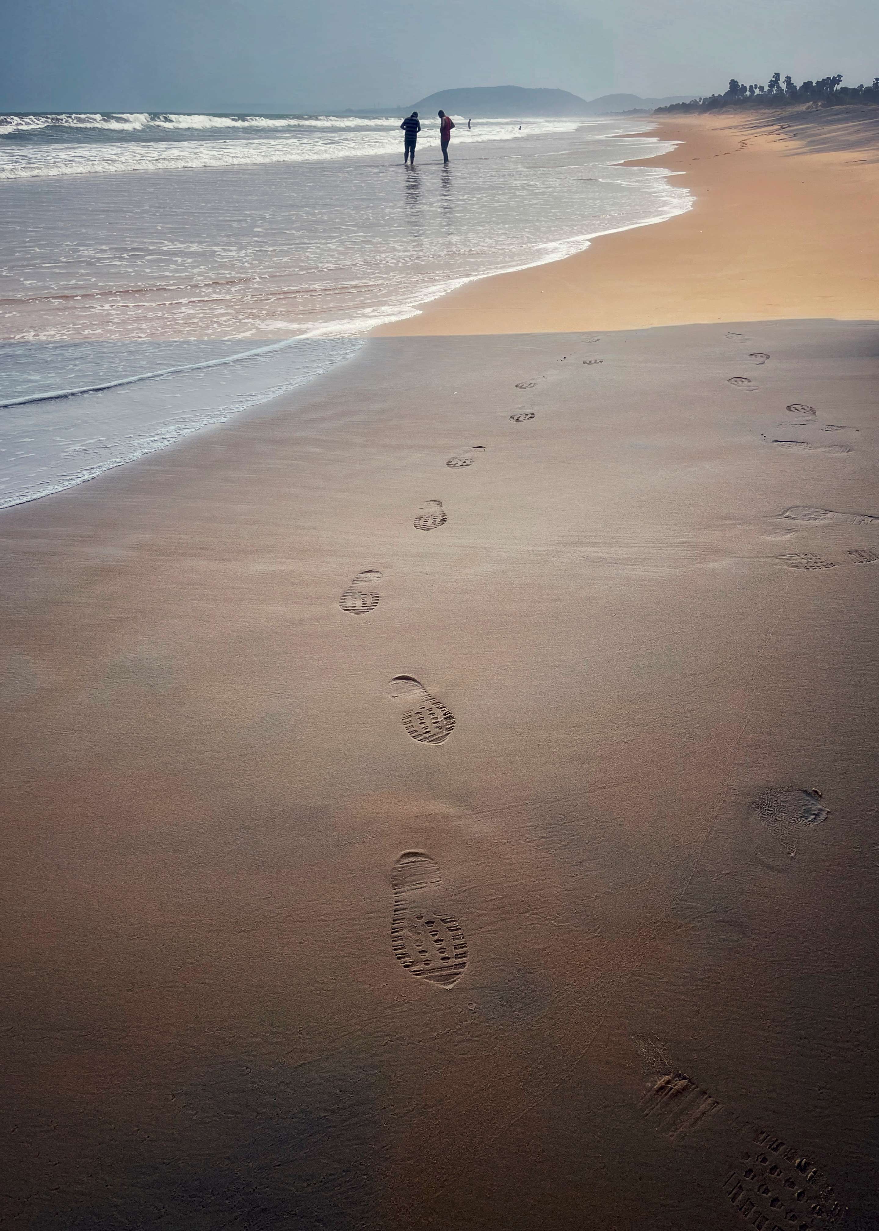 Footprints in sand leading to figures in surf with mountains in background - coastal landscape with human traces