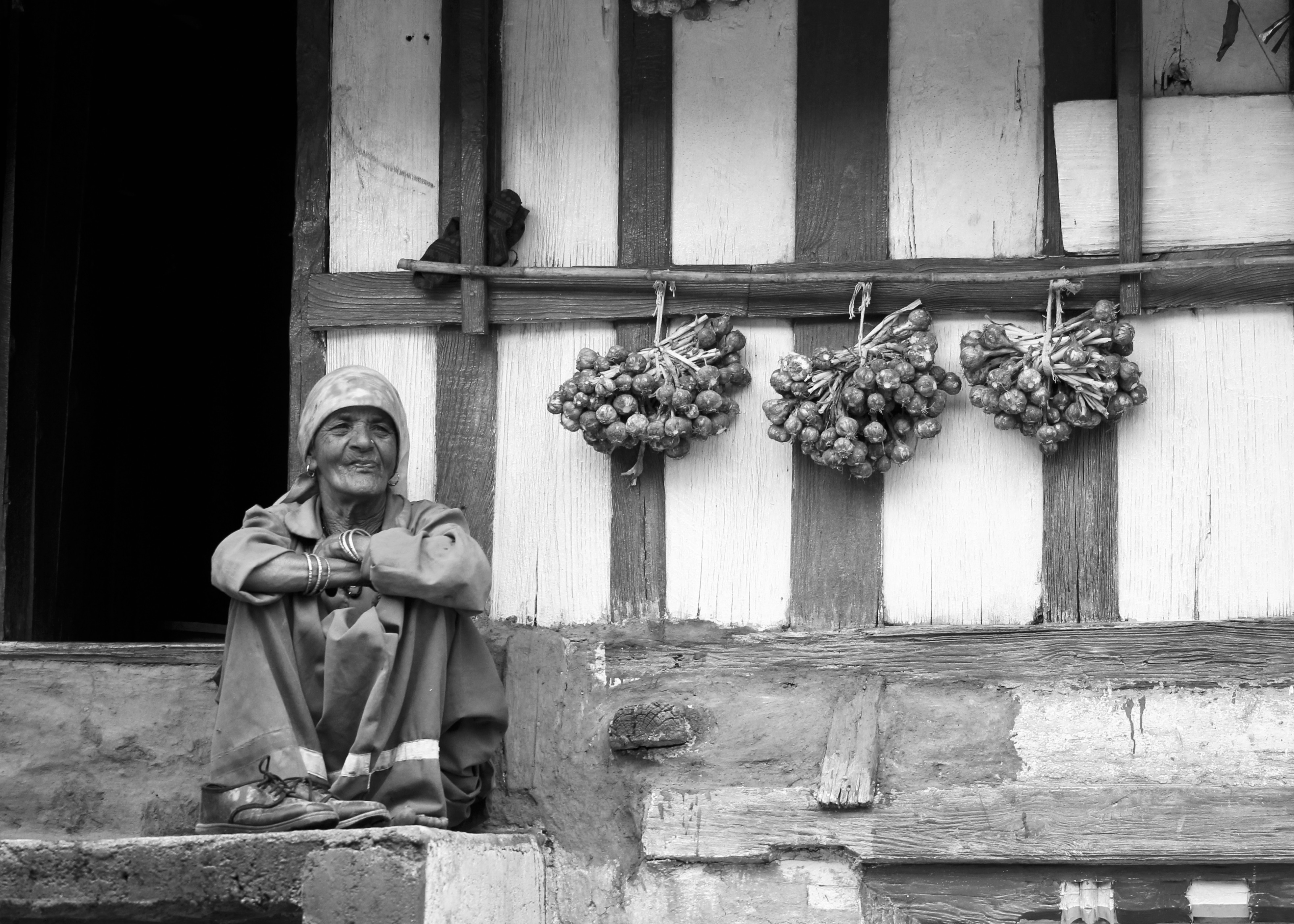 Elderly woman in traditional dress sitting beside hanging vegetables at local market - documentary travel photography