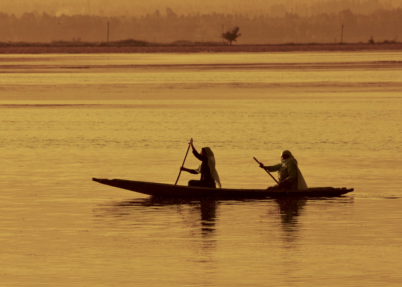 Two people in a small boat on a golden-hued lake, silhouetted against a hazy horizon - Wanderings photography series by Shital Morjaria